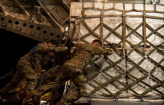 On the left, two U.S. Air Force airmen wearing camouflage push at a pallet that is taller than they are. A third airman is dimly visible behind them, also pushing the pallet from left to right. The pallet is wrapped in a white covering and secured by a cargo net whose brown straps are connected by metal rings.